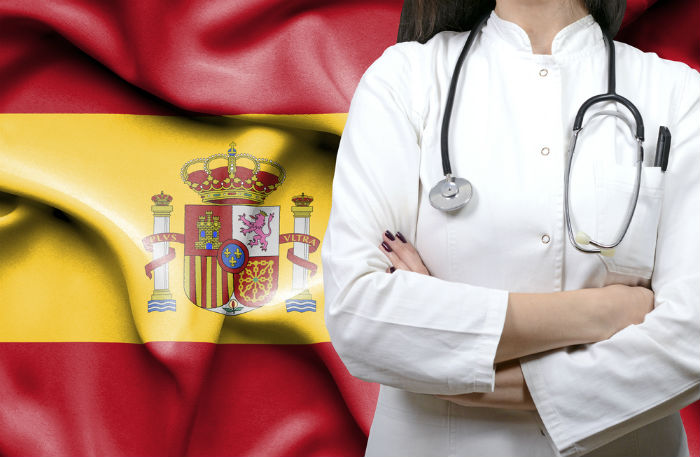White woman photographed from the neck down wearing a white medical jacket and stethoscope standing in front of the Spanish flag.